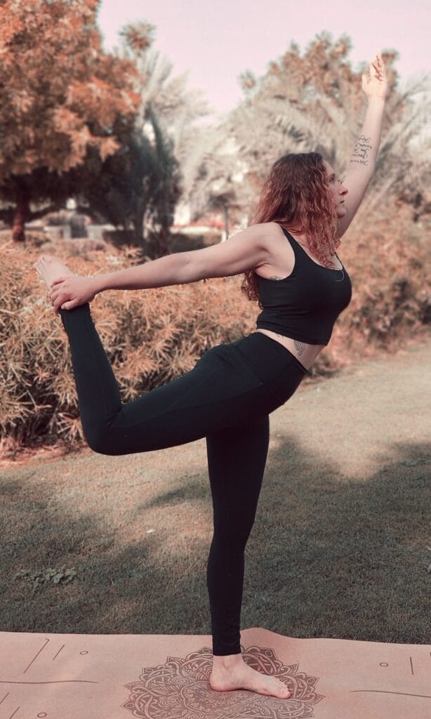 Alina Saibel in Dancer’s Pose (Natarajasana) during an outdoor yoga session – balancing on one leg, other leg lifted and held behind, arm reaching forward, chest open, back arched, gaze focused, embodying grace, strength, and balance in nature
