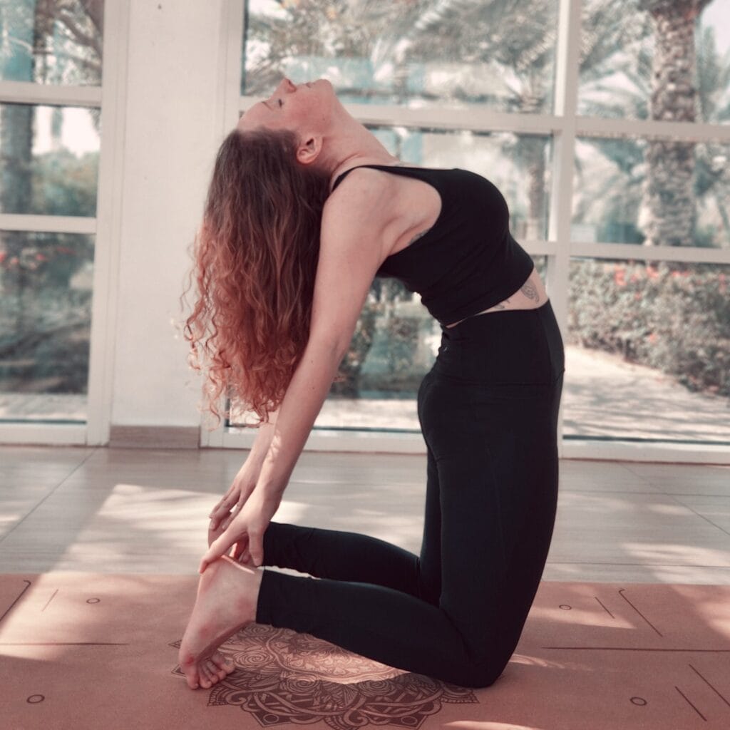Alina Saibel in Camel Pose (Ustrasana) during an outdoor yoga session – kneeling with thighs perpendicular to the ground, hands reaching back to grasp ankles, chest lifted, heart open, head gently dropped back, and a serene sky in the background