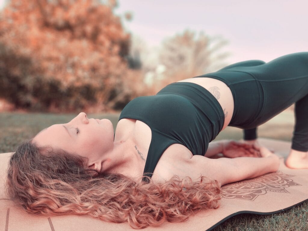 Alina Saibel in Bridge Pose (Setu Bandhasana) during an outdoor yoga session – lying on her back with knees bent, feet grounded, hips lifted, chest open, arms resting on the mat alongside the body, expressing strength, openness, and grounding in nature
