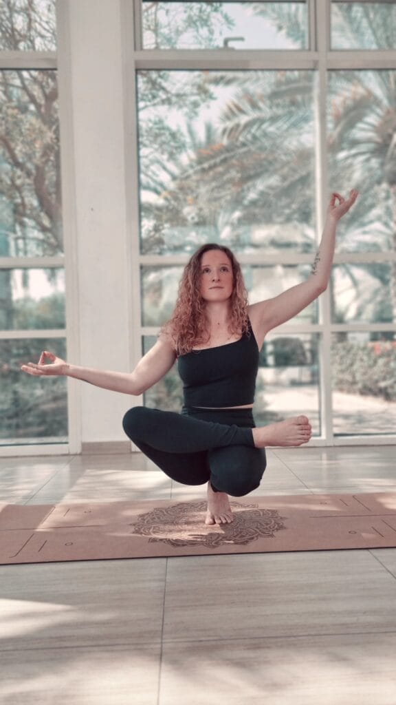 Alina Saibel in Toe Stand (Padangusthasana) during an outdoor yoga session – balancing on the toes of one foot, other foot resting on the thigh in half lotus, gaze soft and focused, embodying balance, stability, and grace