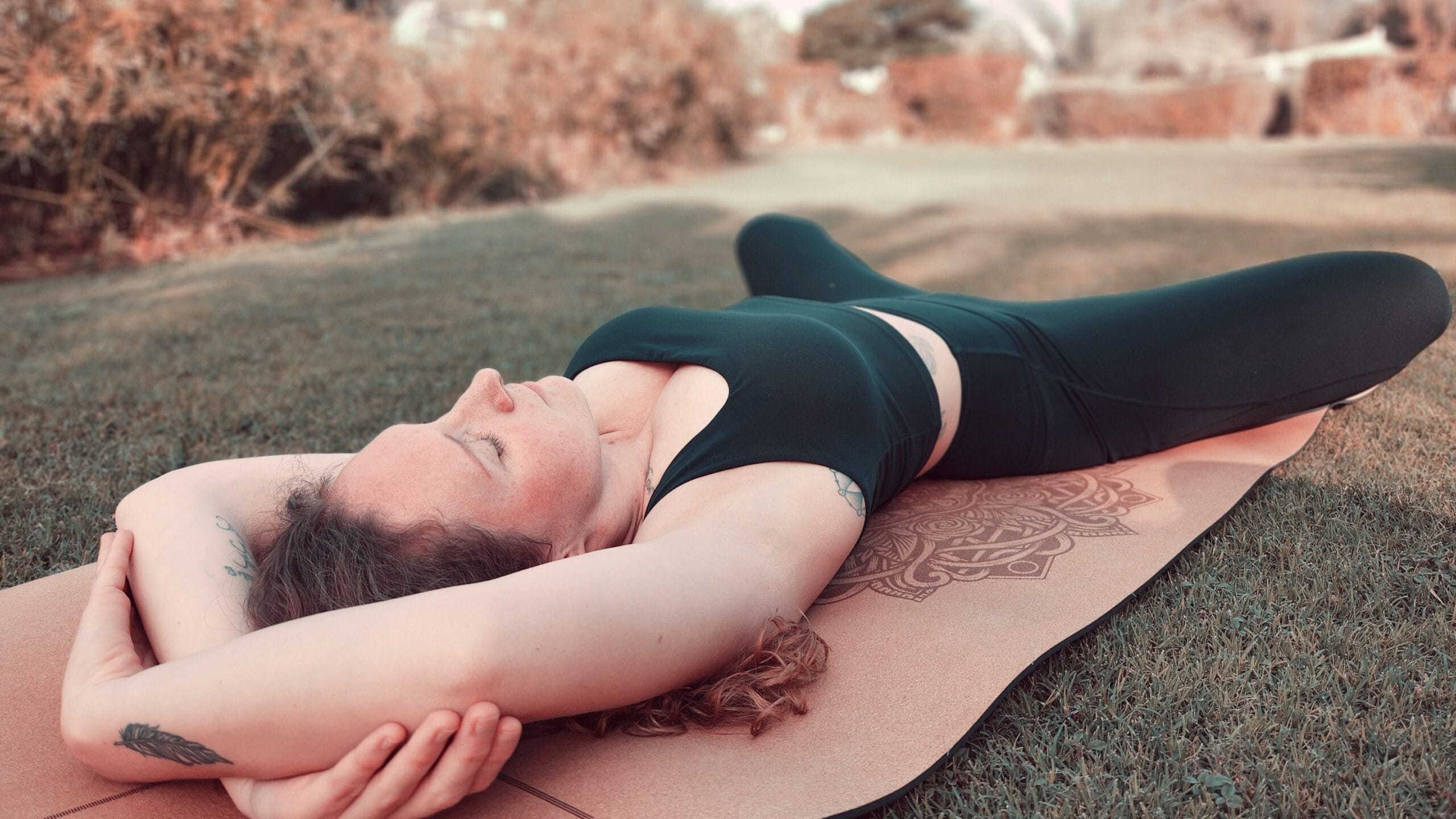 Alina Saibel lying on her back in Reclined Butterfly Pose (Supta Baddha Konasana) during an outdoor yoga session – soles of the feet together, knees gently falling open, hands resting on the belly and heart, eyes closed, radiating calm and relaxation